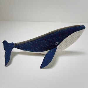 K&ouml;nnte beinhalten: Eine blaue, rei&szlig;verschlussversehene Tasche in Form eines Wals mit wei&szlig;em Bauch und einem roten und blauen gemusterten Stoff. Die Tasche ist aus Stoff und hat einen Rei&szlig;verschluss.