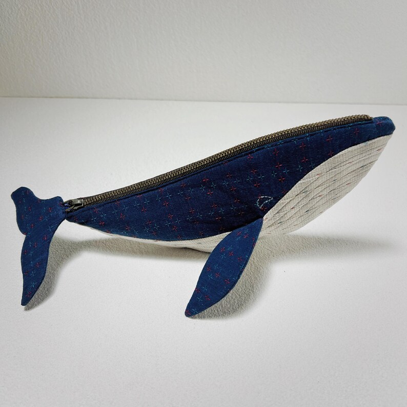 K&ouml;nnte beinhalten: Eine blaue, rei&szlig;verschlussversehene Tasche in Form eines Wals mit wei&szlig;em Bauch und einem roten und blauen gemusterten Stoff. Die Tasche ist aus Stoff und hat einen Rei&szlig;verschluss.