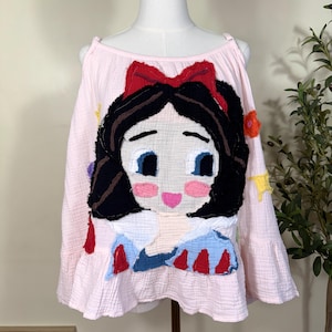 May include: A light pink cold-shoulder top featuring a large, textured fabric appliqué of a character with black hair, a red bow, and a blue and red dress. The garment has long sleeves with ruffled cuffs and a soft, crinkled fabric texture throughout.