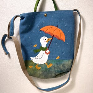 May include: Blue tote bag with a whimsical duck design. The duck is holding an orange umbrella and wearing a green scarf, set against a rainy backdrop. Features a long, adjustable strap and green handles. A charming accessory for everyday use.