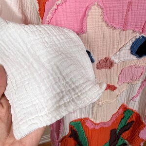 May include: Close-up of a white, textured fabric sleeve. The sleeve is part of a colorful patchwork garment featuring pink, orange, and blue fabric shapes. The garment has a unique, artistic design.