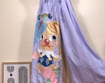 May include: Light purple wide-leg pants with a drawstring waist. The pants feature a large, colorful applique of a cartoon character with blonde hair, blue eyes, and a pink nose. The applique includes blue, pink, and yellow fabric.
