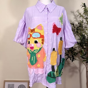 May include: A light purple button-down shirt with a textured fabric finish, featuring a large embroidered patch of a yellow cat wearing goggles and a green scarf, alongside smaller patches of a red butterfly and yellow cattail plants.