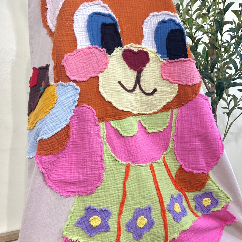 May include: A colorful, handmade, stuffed animal in the shape of a cartoon fox. The fox has orange fur, blue eyes, and a pink and green dress with purple flower accents. The toy is made of layered, textured fabric.