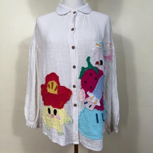 May include: White button-down shirt with long sleeves and a rounded collar. The shirt features colorful, whimsical appliqués of a strawberry, flower, and other cartoon characters. The shirt has wooden buttons.