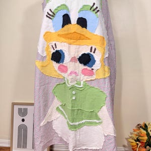 May include: A whimsical, handmade dress featuring a large, colorful applique of a cartoon girl's face. The dress has a white background with a red and white striped pattern. The girl's face has yellow hair, blue eyes, and pink cheeks.