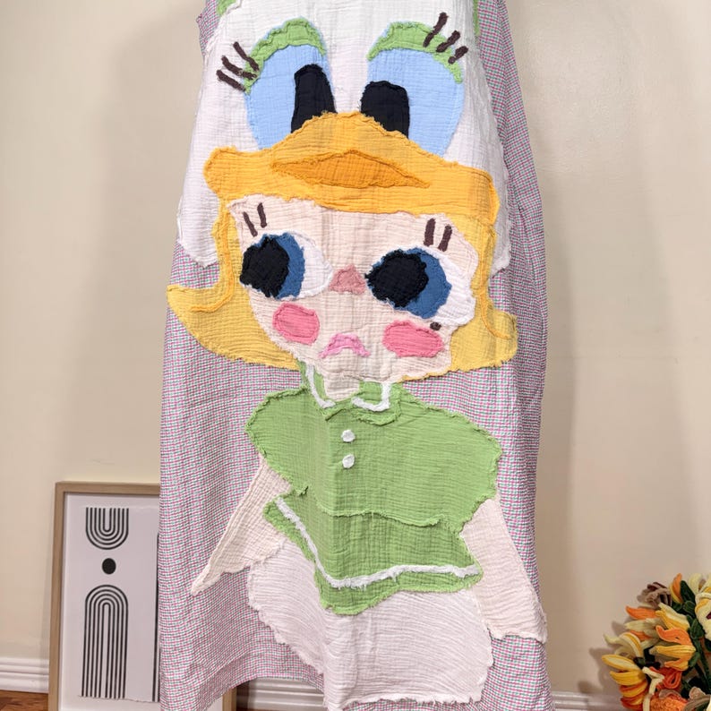 May include: A whimsical, handmade dress featuring a large, colorful applique of a cartoon girl's face. The dress has a white background with a red and white striped pattern. The girl's face has yellow hair, blue eyes, and pink cheeks.