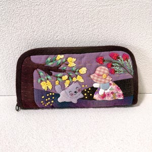 May include: A rectangular fabric wallet with a dark brown border and a purple background. The front features embroidered yellow mimosa flowers on a branch, a small gray cat, and a figure wearing a plaid hat and a floral print dress.