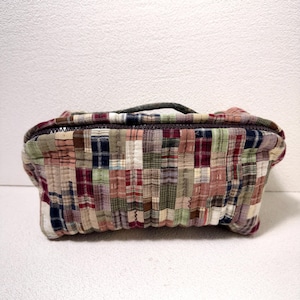May include: A rectangular zippered pouch featuring a quilted patchwork design composed of various plaid fabric squares in muted tones of red, green, blue, brown, and cream. The bag includes a small fabric handle on top and a dark zipper closure.