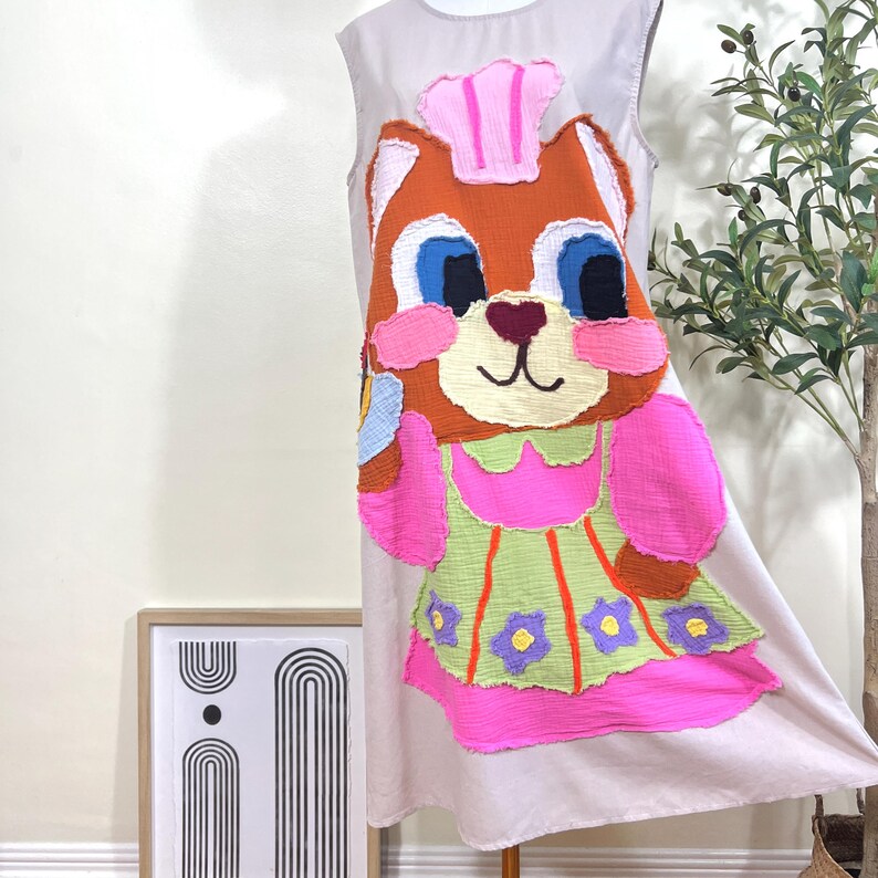 May include: A sleeveless, light pink dress featuring a large, colorful applique of a cartoon squirrel. The squirrel has orange fur, blue eyes, and is wearing a pink chef's hat and apron. The apron is green with pink trim and purple flower accents.