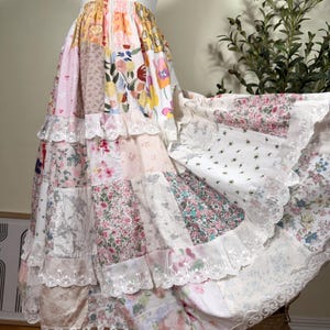May include: A floor-length patchwork skirt featuring a variety of floral and patterned fabric squares in shades of pink, yellow, blue, and white. The skirt has an elastic waistband and multiple tiers accented with white lace trim along the edges.