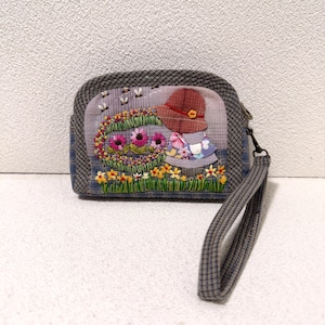 May include: A zippered pouch with a wrist strap, featuring a gray plaid border and a central panel with embroidered flowers, green grass, and a figure wearing a red hat and a floral dress. The design includes small flying insects above the flowers.