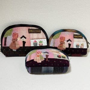 Könnte beinhalten: Drei gequiltete Stoffbeutel mit einem rosafarbenen und braunen Patchwork-Design. Jeder Beutel zeigt eine kleine gestickte Figur und das Wort "Willkommen".