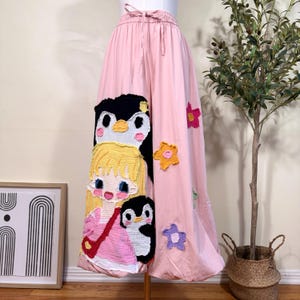 Peut inclure: Pantalon large rose clair avec une taille élastique à cordon de serrage et de grandes appliques en tissu. Le motif représente un personnage blond avec deux manchots et plusieurs patchs en forme d'étoile colorés sur le tissu.