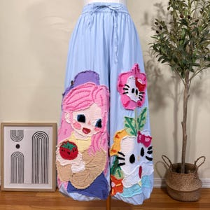 Puede incluir: Pantalones anchos de color azul claro con cintura de cordón y grandes aplicaciones de tela texturizada. La pierna izquierda muestra un personaje de pelo rosa sosteniendo una fresa roja, mientras que la derecha presenta un gato blanco con una flor rosa.