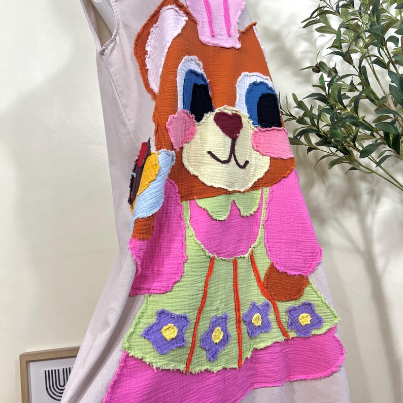 May include: A sleeveless dress featuring a large, colorful applique of a cartoon bear. The bear has orange, pink, blue, and white details. The dress has a pink skirt and a green bodice with purple and yellow flower accents. The dress is made of a soft, textured fabric.