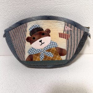 May include: A handmade, oval-shaped pouch featuring a patchwork design. The central panel showcases a brown bear wearing a hat and scarf. The pouch is trimmed with blue and gray patterned fabric, and includes a small flag detail.