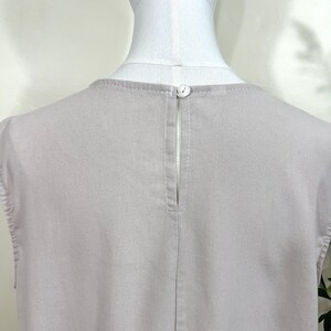 May include: Close-up of a light gray sleeveless top. The back of the top features a small button closure at the neckline and a vertical slit. The fabric has a subtle texture and is neatly stitched.