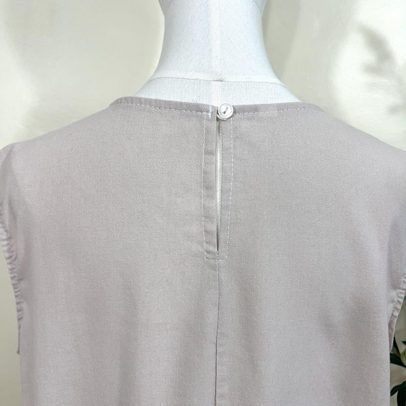May include: Close-up of a light gray sleeveless top. The back of the top features a small button closure at the neckline and a vertical slit. The fabric has a subtle texture and is neatly stitched.