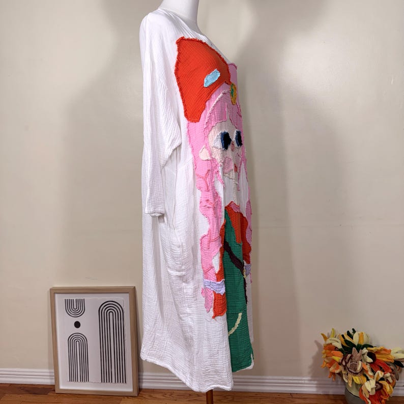 May include: White long-sleeve dress with a colorful applique design. The applique features a cartoon character with a large red bow, pink hair, and green and blue accents. The dress is displayed on a mannequin.