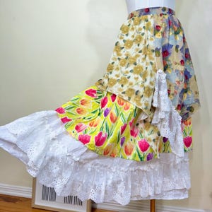 May include: A multi-layered skirt with a patchwork design. The skirt features floral patterns in yellow, pink, and purple, with a white lace trim. The skirt is displayed on a mannequin.