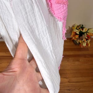 May include: Close-up of a white, textured fabric with a pink, fringed accent. The fabric appears to be a lightweight material, possibly cotton or linen. The pink accent adds a pop of color and texture to the design.
