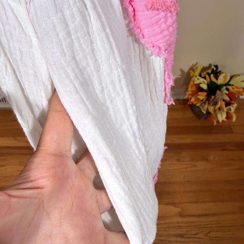 May include: Close-up of a white, textured fabric with a pink, fringed accent. The fabric appears to be a lightweight material, possibly cotton or linen. The pink accent adds a pop of color and texture to the design.