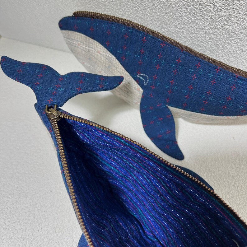 K&ouml;nnte beinhalten: Eine blaue, rei&szlig;verschlussversehene Tasche in Form eines Wals mit einem roten und blauen gemusterten Stoff. Die Tasche ist ge&ouml;ffnet und zeigt ein blaues und gr&uuml;nes gestreiftes Futter.