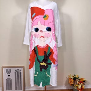 May include: White long-sleeve dress featuring a colorful, textured illustration of a girl with pink hair, a red bow, and green overalls. The girl is holding a brown bunny. The dress has a V-neck and a loose fit.