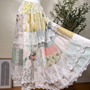 May include: A long, tiered patchwork skirt featuring a variety of pastel-colored fabric panels with floral, geometric, and striped patterns. The skirt is accented with horizontal rows of white scalloped lace trim along the tiered seams and hemline.