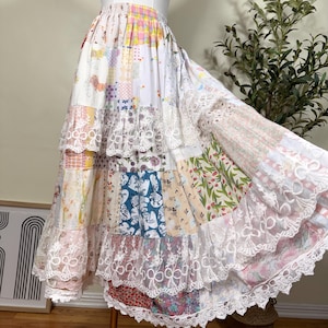 Könnte beinhalten: Ein langer, gestufter Patchwork-Rock mit einer Vielzahl von floralen, karierten und gemusterten Stoffquadraten in Pastell- und leuchtenden Farben. Der Rock hat einen elastischen Bund und ist mit breiter, weißer, bogenförmiger Spitzenborte verziert.