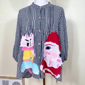 Peut inclure: Un chemisier à carreaux noirs et blancs avec un col mandarin et des boutons en bois. Le chemisier présente des motifs appliqués colorés d'un chat de dessin animé et du Père Noël. Les manches sont aux trois quarts.
