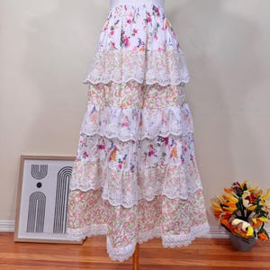 May include: A long, tiered skirt with a floral pattern and lace detailing. The skirt has layers of white fabric adorned with pink, purple, and yellow flowers, and white lace trim. The skirt is displayed on a mannequin.