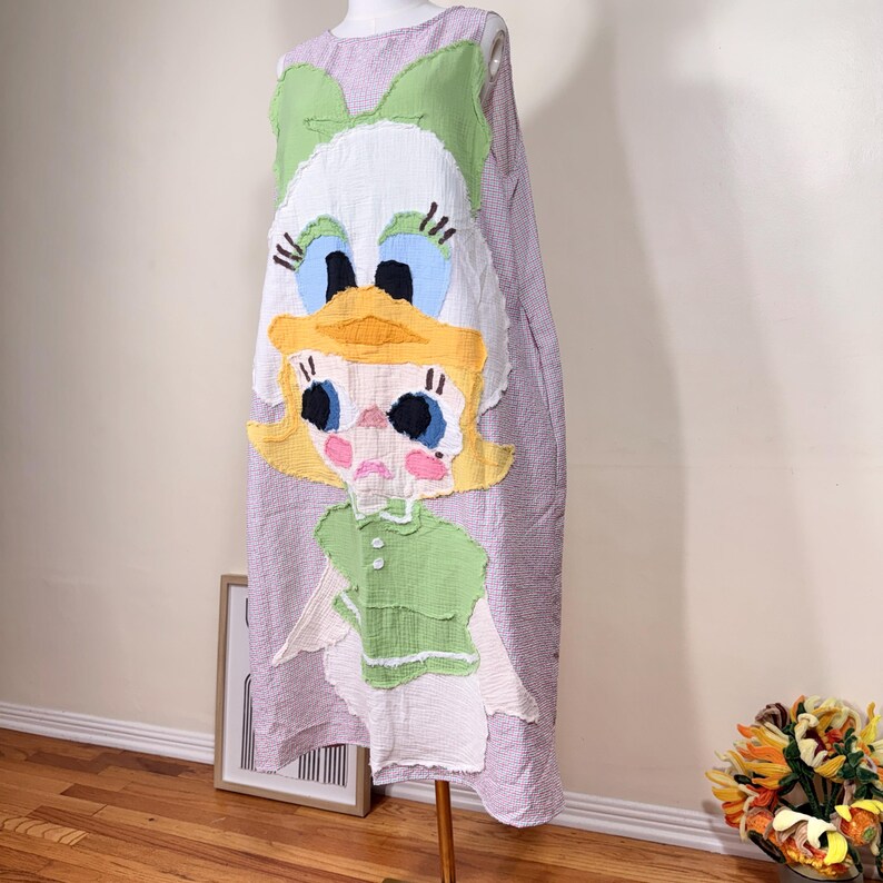 May include: A lavender and white patterned dress featuring a large, colorful cartoon character design. The character has a green bow, yellow hair, blue eyes, and a green top. The dress is displayed on a mannequin.