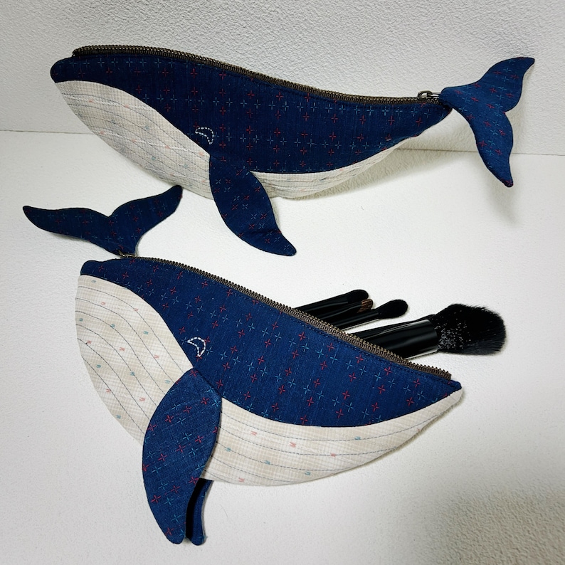 K&ouml;nnte beinhalten: Zwei blaue, walf&ouml;rmige Rei&szlig;verschlusstaschen aus Stoff mit wei&szlig;em und blauem Muster. Die Tasche unten hat einen offenen Rei&szlig;verschluss und Make-up-Pinsel befinden sich darin.