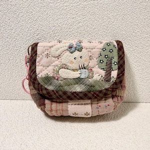 Pode incluir: Uma bolsa crossbody acolchoada com um design de patchwork. A bolsa apresenta um coelho aplicado com um laço azul e uma árvore. A bolsa tem uma alça rosa e um fecho de aba. A bolsa é feita de vários tecidos em tons de rosa, verde e castanho.