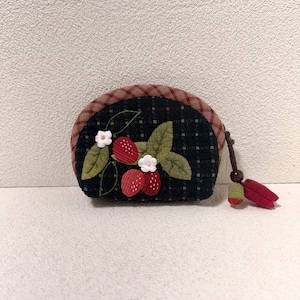 Puede incluir: Un pequeño monedero redondo con un fondo negro con un diseño de fresas. Las fresas son rojas con puntos blancos y las hojas son verdes. La parte superior del bolso es de cuadros marrones y tiene una borla marrón con una cuenta de madera.