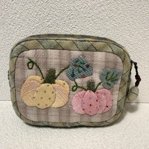May include: A rectangular fabric pouch featuring a quilted olive green and brown plaid border. The center panel displays two appliquéd pumpkins, one in yellow with polka dots and one in pink with small squares, accented by embroidered green leaves.