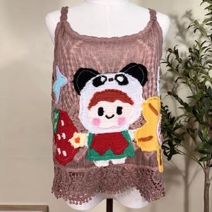 May include: A mauve sleeveless top with a textured fabric and a scalloped lace hem. The front features a large, colorful fabric appliqué of a character wearing a panda hood, flanked by a red strawberry shape and a yellow leaf-like design.