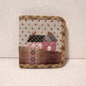 May include: A quilted, square-shaped item with a house design. The top section features a light beige fabric with small, colorful squares. Below, a brown fabric with a floral pattern. The house has a black and red roof and pink walls.