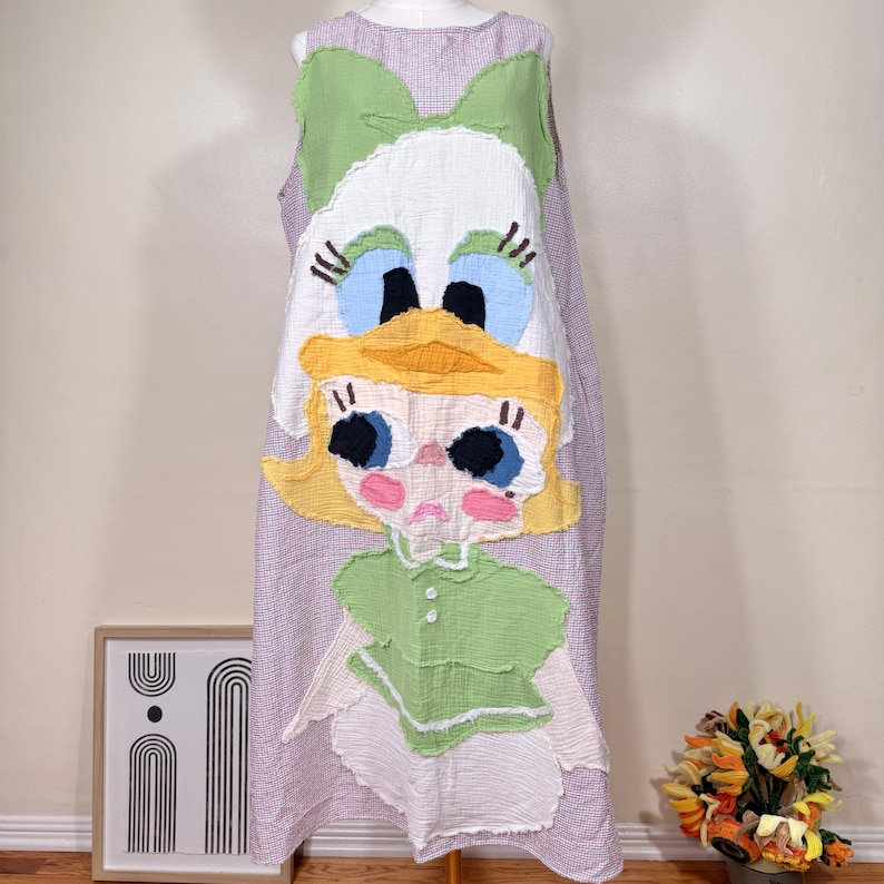 May include: A sleeveless, lilac and white checkered dress featuring a large, whimsical Daisy Duck applique. The applique includes a green bow, white head, blue eyes, yellow beak, and a green top. The dress is displayed on a mannequin.