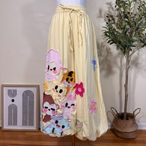 Puede incluir: Un pantalón de pierna ancha en color amarillo pálido con cintura elástica y cordón ajustable. La parte delantera está decorada con varios parches de tela texturizada y colorida que representan caras de animales y flores pequeñas.