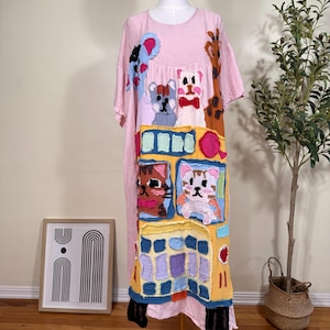 May include: A light pink short-sleeved dress featuring a large, colorful patchwork design on the front. The applique includes various animal faces, including cats and a giraffe, alongside geometric shapes in blue, yellow, and purple fabric textures.