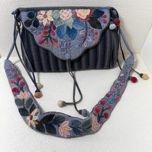 May include: A denim blue crossbody bag with a floral applique design. The bag features a flap closure and a long, embroidered strap. The floral design includes pink, yellow, and blue flowers with green leaves. The bag has a striped pattern.