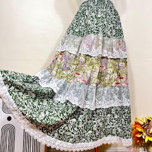 May include: A tiered maxi skirt with a floral pattern in shades of green, purple, and white. The skirt has lace trim between each tier. The skirt is made of a lightweight fabric, suitable for a bohemian or vintage-inspired style.