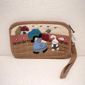 Puede incluir: Un bolso de mano acolchado con una escena caprichosa con una niña, un conejo y una casa. El bolso tiene un borde a cuadros marrón y una correa de muñeca. La escena incluye una niña con un vestido azul, un conejo blanco y una cereza roja.