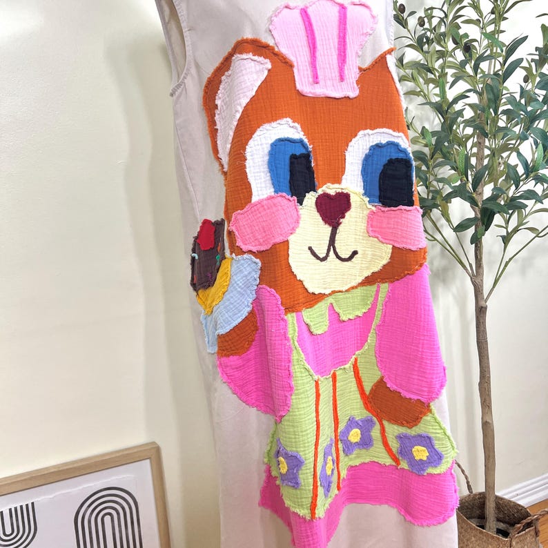 May include: A beige sleeveless dress featuring a large, colorful applique of a cartoon cat chef. The cat is orange with pink cheeks, blue eyes, and a pink chef's hat. The dress has a pink and green apron with purple flower accents.