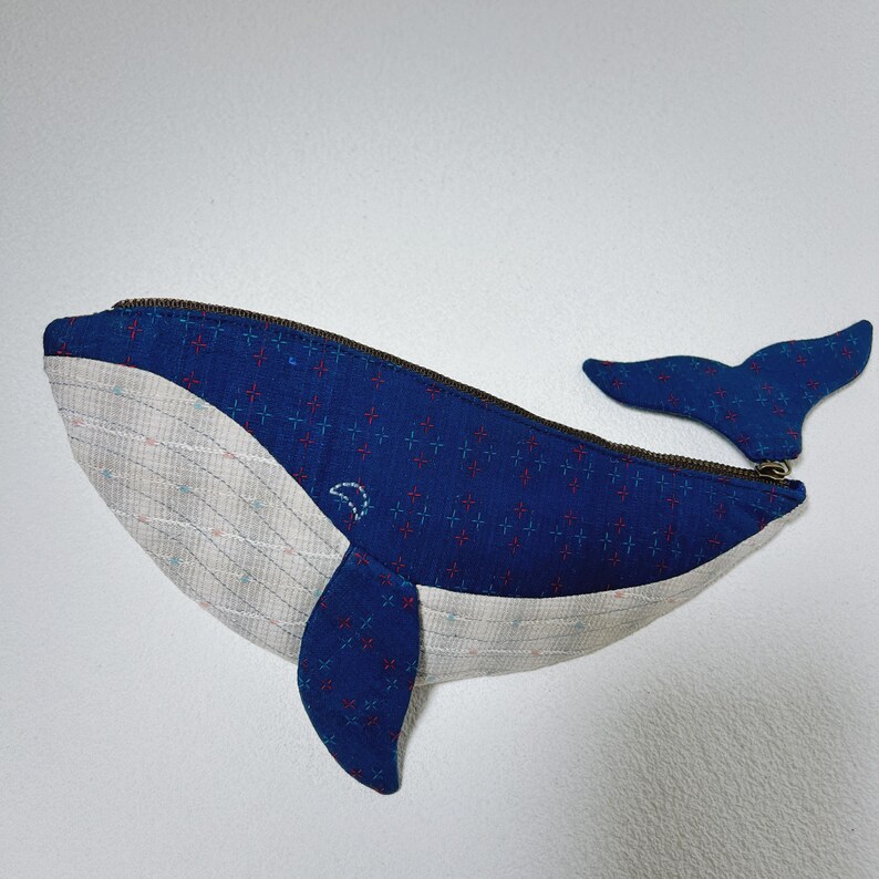 K&ouml;nnte beinhalten: Eine blaue, walf&ouml;rmige Rei&szlig;verschlusstasche aus einem wei&szlig;en und blauen gemusterten Stoff. Die Tasche ist aus Stoff und hat einen Rei&szlig;verschluss.