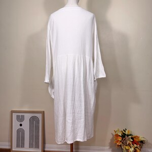 May include: A white long-sleeve dress with a textured, crinkled fabric. The dress has a V-neck and a gathered waist, creating a flowing silhouette. The dress is displayed on a mannequin, showcasing its design and fit.