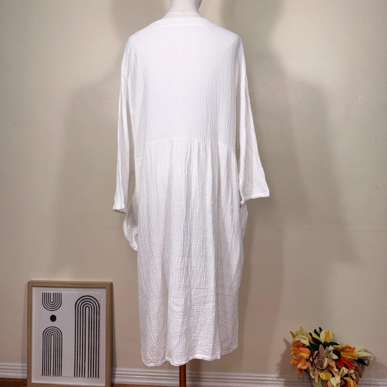 May include: A white long-sleeve dress with a textured, crinkled fabric. The dress has a V-neck and a gathered waist, creating a flowing silhouette. The dress is displayed on a mannequin, showcasing its design and fit.